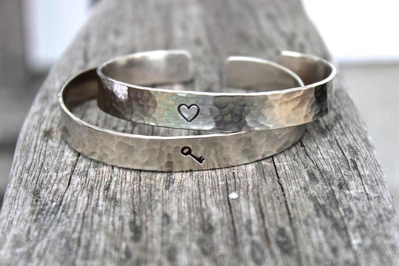 May include: Two silver-colored hammered metal cuff bracelets. One bracelet has a heart symbol, and the other has a key symbol. The bracelets are displayed on a weathered wooden surface.