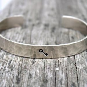 May include: Silver cuff bracelet with a hammered texture and a small key symbol. The open-ended bracelet is displayed on a weathered wooden surface, showcasing its simple, elegant design. A piece of jewelry.