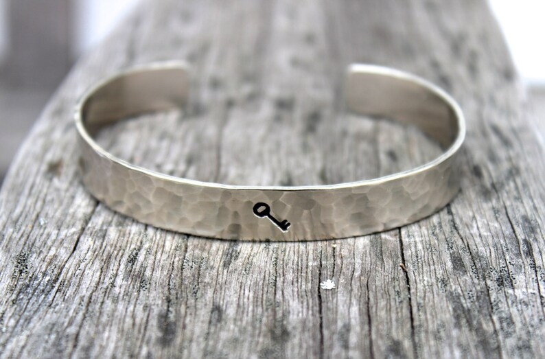May include: Silver cuff bracelet with a hammered texture and a small key symbol. The open-ended bracelet is displayed on a weathered wooden surface, showcasing its simple, elegant design. A piece of jewelry.