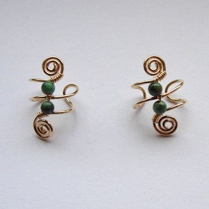 May include: Two gold-colored ear cuffs, each featuring a spiral design at the top and bottom, connected by a curved wire. Each cuff has two small, round, dark green beads. The ear cuffs are displayed on a light background.