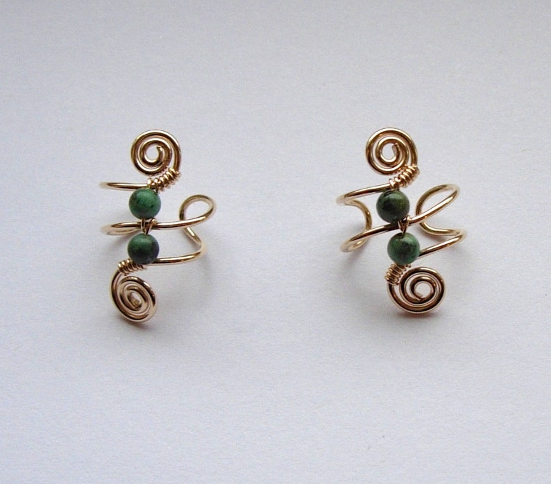 May include: Two gold-colored ear cuffs, each featuring a spiral design at the top and bottom, connected by a curved wire. Each cuff has two small, round, dark green beads. The ear cuffs are displayed on a light background.