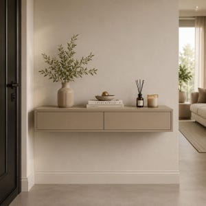 Op de afbeelding: Een zwevende, taupekleurige console met twee lades, versierd met een vaas met groen, boeken, een kom met een gouden bol, een diffuser en een kaars. De console is gemonteerd op een muur in neutrale tinten.