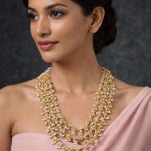 Peut inclure: Un collier doré à plusieurs rangs composé de travail métallique complexe et de nombreuses petites perles blanches. Le design se compose de plusieurs brins en cascade créant une apparence dense et texturée, reposant sur la zone de la clavicule.