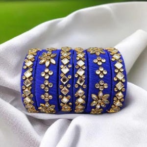 May include: A set of six blue bangles with gold-colored floral and geometric embellishments. The bangles are resting on a white cloth, with a blurred green background.