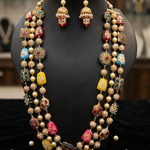 Puede incluir: Un conjunto de collar y pendientes dorados, exhibido en un maniquí negro. El collar presenta múltiples hilos de perlas de imitación y colgantes coloridos y ornamentados en rojo, amarillo, azul y verde. Los pendientes tienen un diseño a juego.