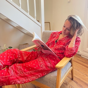 May include: A person is wearing a red paisley pajama set, comprising a shirt and trousers, while seated in a grey armchair. The pajamas have white trim and a bandana pattern. The person is reading a magazine.