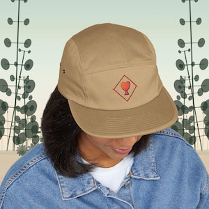 Puede incluir: Una gorra de cinco paneles de color beige con un pequeño diamante rojo bordado que contiene un icono de corazón con degradado naranja y rojo en la parte frontal. La gorra tiene una visera plana y ojales metálicos en los laterales.