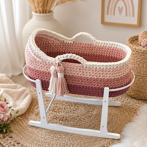 Könnte beinhalten: Ein gehäkelter Babykorb mit einem Farbverlauf in Rosa und Burgunderrot. Der Korb verfügt über zwei integrierte Griffe, eine dekorative Quaste und wird von einem weißen Schaukelgestell aus Holz mit geschwungenem Rahmen getragen.