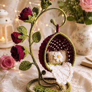 May include: A crochet hanging chair ornament featuring a deep red, lattice-patterned egg-shaped seat suspended from a green vine-like stand adorned with dark red rose blossoms and green leaves, containing a small beige pillow with a white daisy.
