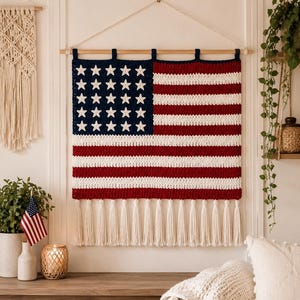 Puede incluir: Un tapiz rectangular de ganchillo con el diseño de la bandera estadounidense, que presenta un cantón azul oscuro con estrellas blancas, rayas horizontales rojas y blancas alternas y una fila de largos flecos blancos en el borde inferior.