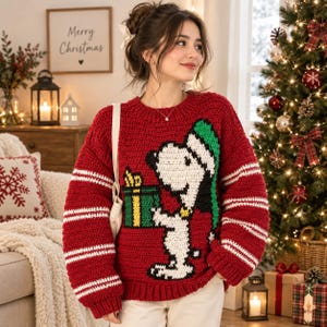 May include: A red long-sleeve crochet sweater featuring a white Snoopy character wearing a green hat and holding a green gift box with a yellow ribbon. The sweater has white horizontal stripes on the sleeves and a ribbed crew neckline.