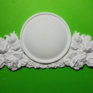 Può includere: Una placca da parete decorativa bianca con un'area circolare centrale e accenti floreali di rose. La placca è realizzata in un materiale solido ed è posta su uno sfondo verde.