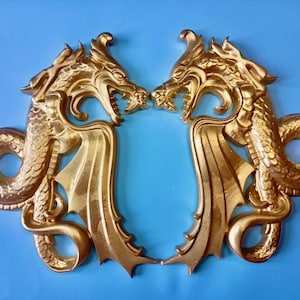 Könnte beinhalten: Zwei goldfarbene Drachen-Wandskulpturen, die sich gegenüberstehen. Jeder Drache hat detaillierte Schuppen, Flügel und einen offenen Mund. Der Hintergrund ist einfarbig hellblau.