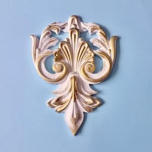 Può includere: Applique da parete decorativo bianco e oro con un design simmetrico. Il pezzo ornato presenta un elemento verticale centrale con accenti a spirale e uno sfondo azzurro.
