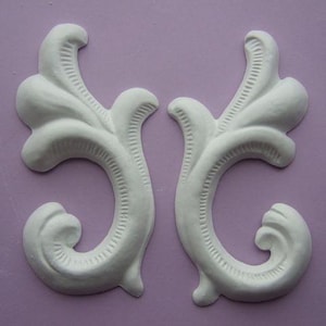 Può includere: Due elementi decorativi bianchi, ornati e simmetrici. Ogni elemento ha un design curvo e aperto con forme strutturate a forma di foglia e un ricciolo. Lo sfondo è di colore lilla chiaro.