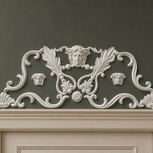 Op de afbeelding: Een wit decoratief architectonisch fronton met een centraal reliëf van een menselijk gezicht omringd door ingewikkelde acanthusbladkrullen en twee kleinere gezichtsmotieven aan weerszijden, gemonteerd boven een lichtgekleurd rechthoekig deurkozijn.