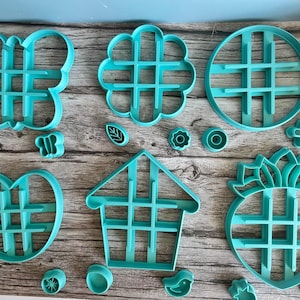 May include: A collection of teal plastic cookie cutters in various shapes including a butterfly, flower, circle, heart, house, and pineapple, each featuring an internal grid structure, accompanied by several smaller matching mini cutters.