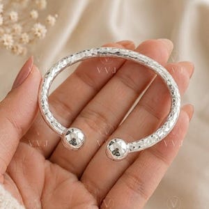 May include: A silver open cuff bracelet with a hammered texture. The bracelet has a rounded shape with two spherical end caps. The jewellery is displayed on a neutral background.