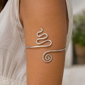 May include: A silver-toned arm cuff featuring a continuous wire design that forms a series of undulating waves and a circular spiral at one end. The metal band wraps around the upper arm, showcasing a smooth, polished finish and a flexible structure.