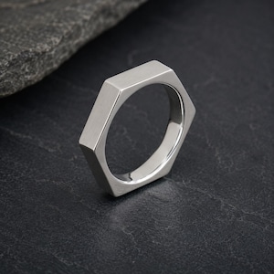 May include: A silver-coloured metal ring featuring a distinct hexagonal outer shape. The band has a smooth, brushed finish on its flat exterior surfaces, while the inner circumference is polished and rounded to form a circular opening for the finger.