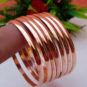 Puede incluir: Una pila de brazaletes de color cobre. El metal liso y pulido refleja la luz, creando reflejos y sombras. Los brazaletes están apilados, mostrando su diseño simple y elegante. El fondo presenta follaje verde y una flor roja.