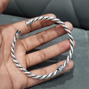 Può includere: Un bracciale rigido in metallo color argento caratterizzato da un design a corda intrecciata. Il bracciale presenta uno stile aperto con due tappi cilindrici alle estremità, ciascuno dettagliato con sottili scanalature orizzontali.
