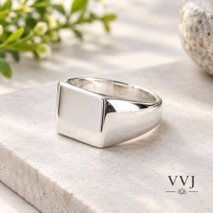 Könnte beinhalten: Ein polierter Siegelring aus Silber mit einer glatten, flachen, rechteckigen Oberfläche und einer breiten, massiven Ringschiene. Der Ring hat eine reflektierende metallische Oberfläche und ist mit dem Text VVJ und einer Lotusblüte markiert.