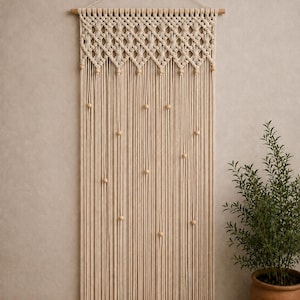 Pode incluir: Uma tapeçaria de macramé cor de creme suspensa por um bastão de madeira com um laço de corda. A secção superior apresenta um padrão complexo de nós em forma de diamante com contas de madeira, e a inferior consiste em longas franjas verticais.