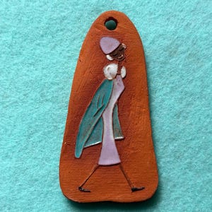 Pode incluir: Pingente cor de terracota com uma figura estilizada a andar. A figura é representada em branco, lavanda e azul-petróleo, usando chapéu e casaco. O pingente tem um furo na parte superior para pendurar.
