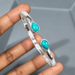 May include: A silver cuff bracelet featuring an open design with two teardrop-shaped turquoise stones at each end. The polished metal band has a smooth, reflective surface, and the bright blue stones display natural dark veining throughout.