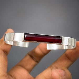 May include: A silver cuff bracelet featuring a smooth, rectangular, deep red stone inlay centred on the band. The metal has a polished finish, and the bracelet is an open-ended design, showcasing a clean, metallic structure with a dark red accent.