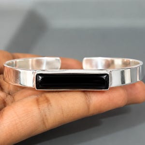 Puede incluir: Un brazalete de plata con una incrustación de piedra negra rectangular y lisa centrada en la parte frontal. La banda de metal tiene un acabado pulido y reflectante con un diseño abierto que permite el ajuste. La piedra negra está engastada.
