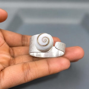 以下が含まれることがあります： テクスチャ加工されたバンドと、中央に茶色の渦巻き模様が入った円形の白い貝殻のインレイが特徴の、調節可能なシルバー調のリング。金属表面はマットなつや消し仕上げで、バンドの背面は開いたデザインになっています。