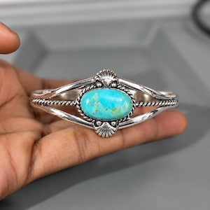 May include: A silver cuff bracelet featuring a central oval turquoise stone with brown matrix. The metal band has a twisted rope texture and decorative shell-like accents at the top and bottom of the stone setting, creating a detailed design.