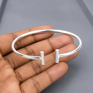 May include: A silver-coloured, open-ended bangle bracelet. The bracelet has a simple, modern design with two vertical bar accents. The bracelet is held in a hand against a grey background. The bracelet is made of a shiny metal.
