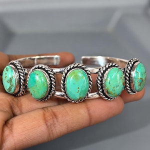 May include: A silver cuff bracelet featuring five oval-shaped turquoise stones. Each stone is set in a polished silver bezel surrounded by a twisted rope-style silver border. The bracelet has a double-band design that connects at the ends.