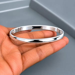 May include: A polished silver bangle bracelet. The bracelet is round and smooth, reflecting light. The bangle is held in a hand against a grey background. This simple, elegant bracelet is a classic jewellery piece.