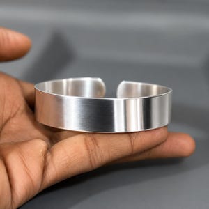 Puede incluir: Una pulsera tipo brazalete ancha y abierta con un acabado plateado cepillado y liso. La pulsera presenta un diseño de banda simple y minimalista con una superficie metálica pulida que refleja la luz, creando un aspecto limpio y uniforme.