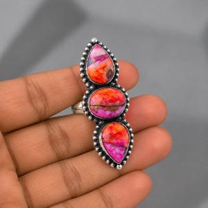 May include: A sterling silver ring featuring three vertically aligned stones with vibrant orange and pink marbled patterns. The stones are set in a beaded silver frame, with a teardrop shape at the top and bottom and an oval shape in the center.