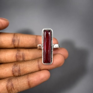 以下が含まれることがあります： 滑らかな金属製のベゼルに垂直にセットされた、長く長方形の深い赤色の石が特徴のシルバーリング。バンドは磨かれたシルバーで作られており、細長い濃いクリムゾン色の宝石がリングの正面中央にしっかりと固定されています。