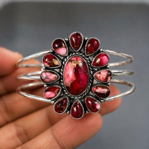 May include: A silver cuff bracelet featuring a large oval centre stone surrounded by twelve smaller teardrop-shaped stones. The stones are a vibrant mix of deep pink, magenta, and dark red with metallic copper veining. The band consists of three thin silver wires.