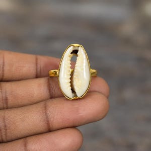 Könnte beinhalten: Ein goldfarbener Ring mit einer mittig platzierten, ovalen weißen Kaurimuschel. Die Muschel ist vertikal in einen glatten, metallisch goldenen Rahmen eingefasst, der mit einem dünnen, kreisförmigen Band verbunden ist und einen Kontrast bildet.