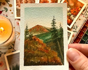 Puede incluir: Una pequeña pintura rectangular de acuarela sobre papel texturizado que representa un paisaje montañoso en otoño. La escena presenta árboles de hoja perenne altos en primer plano, un pico de montaña central y un cielo azul claro. El follaje muestra tonos naranja, rojo y verde oscuro.
