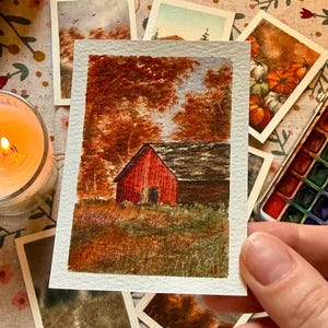 Könnte beinhalten: Ein rechteckiges Aquarellbild auf strukturiertem Papier, das eine rote Scheune umgeben von herbstlichen Bäumen mit orangefarbenem und braunem Laub zeigt. Die Szene umfasst ein grasbewachsenes Feld im Vordergrund und einen bewölkten Himmel.