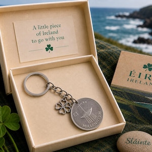 Pode incluir: Um porta-chaves de metal prateado com um pendente de moeda circular marcado com o texto £1 MILLENNIUM e um trevo de quatro folhas, apresentado numa caixa de cartão bege com o texto A little piece of Ireland to go with you.