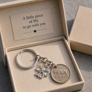 May include: A silver-toned keychain featuring a circular 5P coin charm with a bull design and a four-leaf clover charm, presented in a beige gift box. The box lid interior displays the printed text: A little piece of life to go with you.