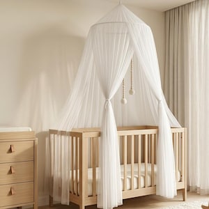 May include: A light wood cot with a white sheer canopy draped from a circular frame suspended above. The canopy features delicate white fabric panels gathered at the sides and includes a hanging mobile with small wooden beads inside.