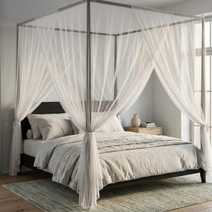 Op de afbeelding: Een hemelbed met een donker frame, voorzien van doorschijnende witte gordijnen die vanaf de bovenste rails naar beneden vallen. Het bed is opgemaakt met lichtgekleurd beddengoed en meerdere kussens onder het frame met dunne verticale steunen.