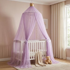 May include: A light purple tulle bed canopy featuring a gold crown topper and integrated string lights. The canopy drapes over a white wooden crib, which contains a beige blanket and a small brown teddy bear sitting on the floor nearby.