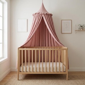 May include: A light wood crib with vertical slats and a white mattress, positioned beneath a dusty rose fabric canopy. The canopy features a gathered ruffle at the top and drapes down in long, soft folds behind the crib, creating a decorative accent.